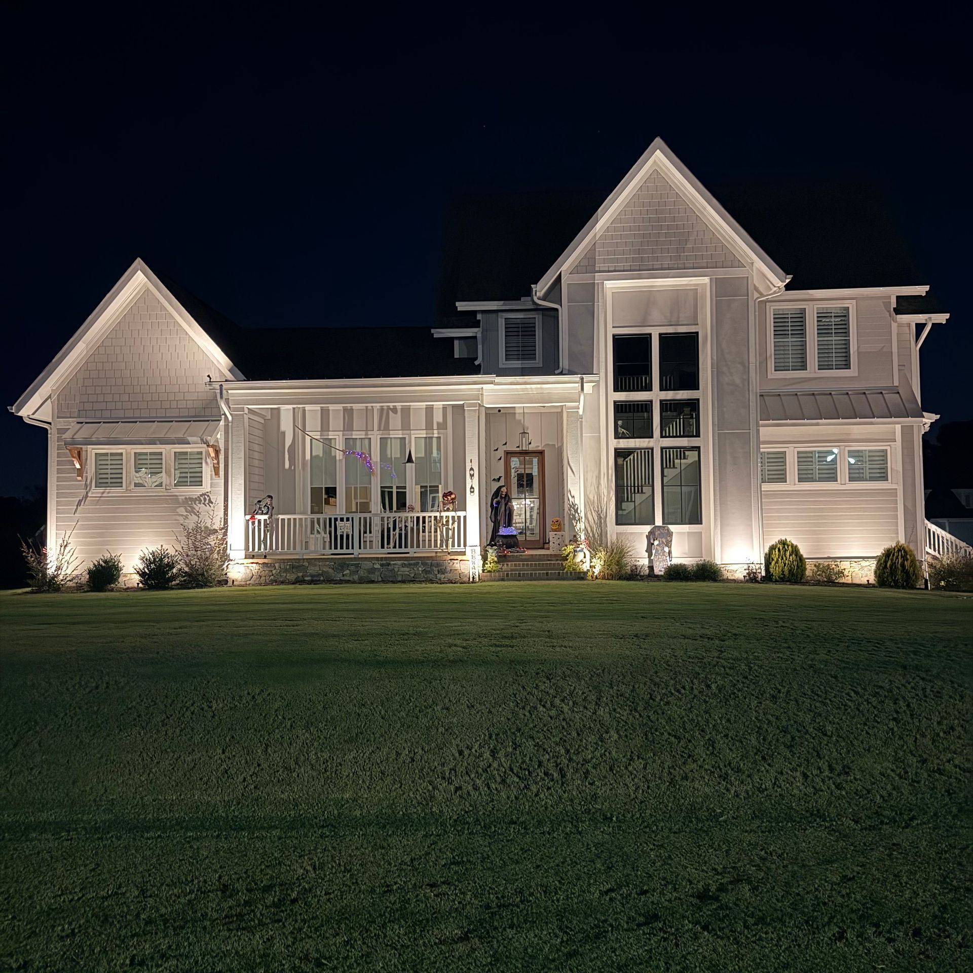 Lit up white house at night with large windows and a porch. Green lawn in the foreground.