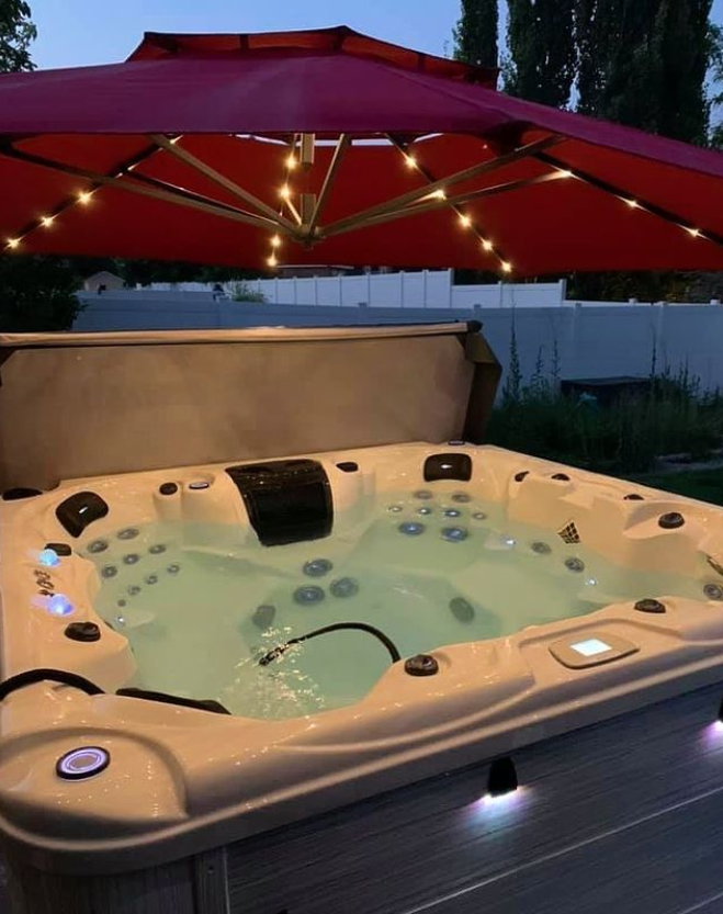 Lit hot tub under a red patio umbrella at dusk, with soft outdoor lighting.
