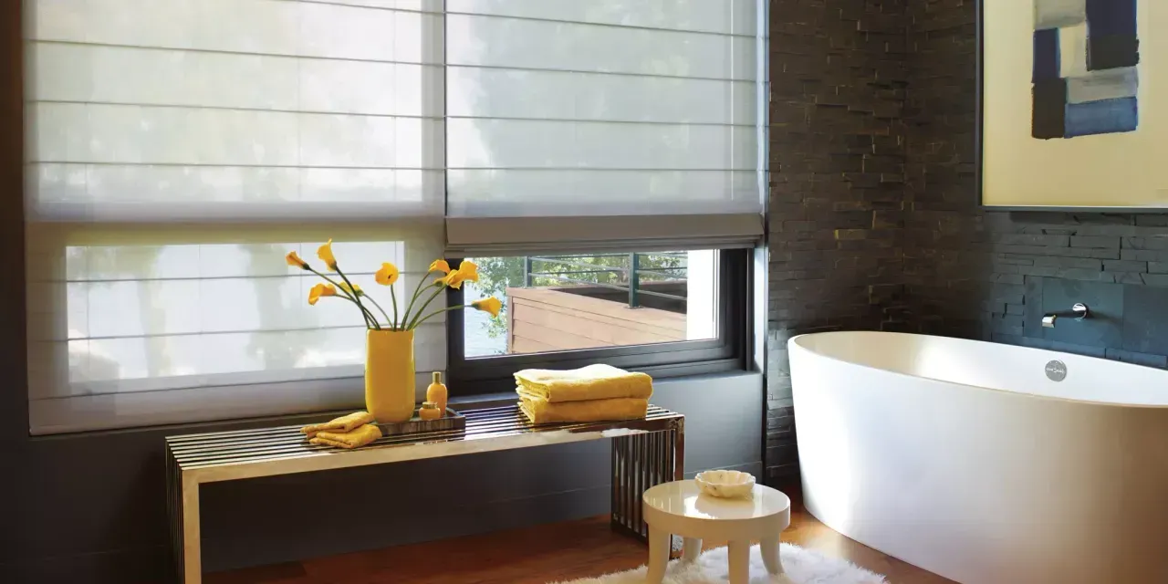 A modern bathroom with a white bathtub, dark tile walls, and sheer window shades. A bench holds flowers and towels.