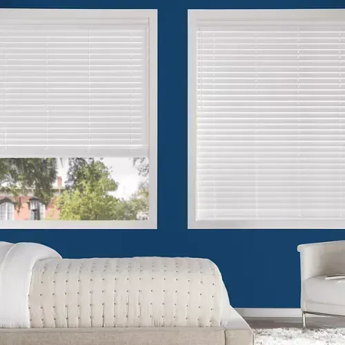 A bedroom with white blinds, a bed, a white chair, and blue walls. Sunlight filters through the blinds.