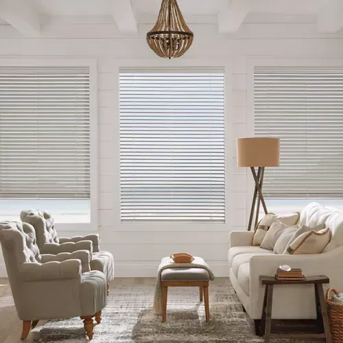 A bright living room with white walls and three windows with blinds. Two armchairs and a sofa face the windows, with a small table and rug.