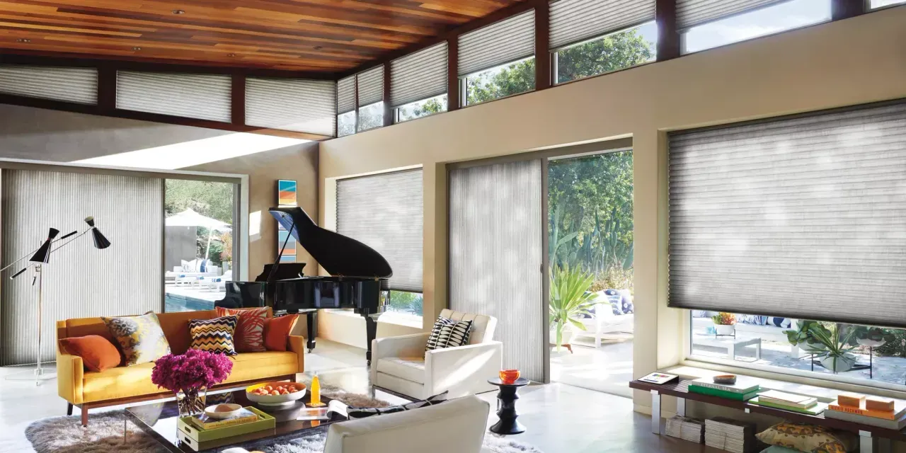 A brightly lit modern living room with a grand piano, sofa, and large windows with shades. Warm wood ceiling and neutral-toned walls.