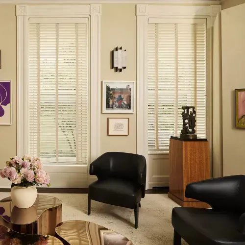 A living room with black leather armchairs, art on the walls, and light-colored window blinds. A vase of pink flowers sits on a gold coffee table.