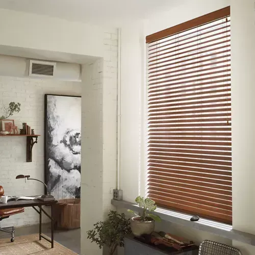 Wooden blinds cover a window in a room with a workspace. Neutral walls surround the window with a glimpse of a desk, art, and plants.