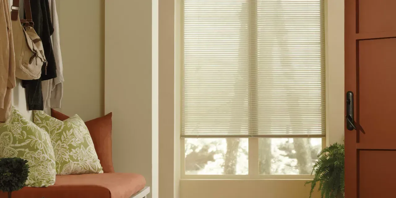 An entryway with a red bench and pillows, a closed door, and a window with blinds. Coats and bags hang on hooks to the left.