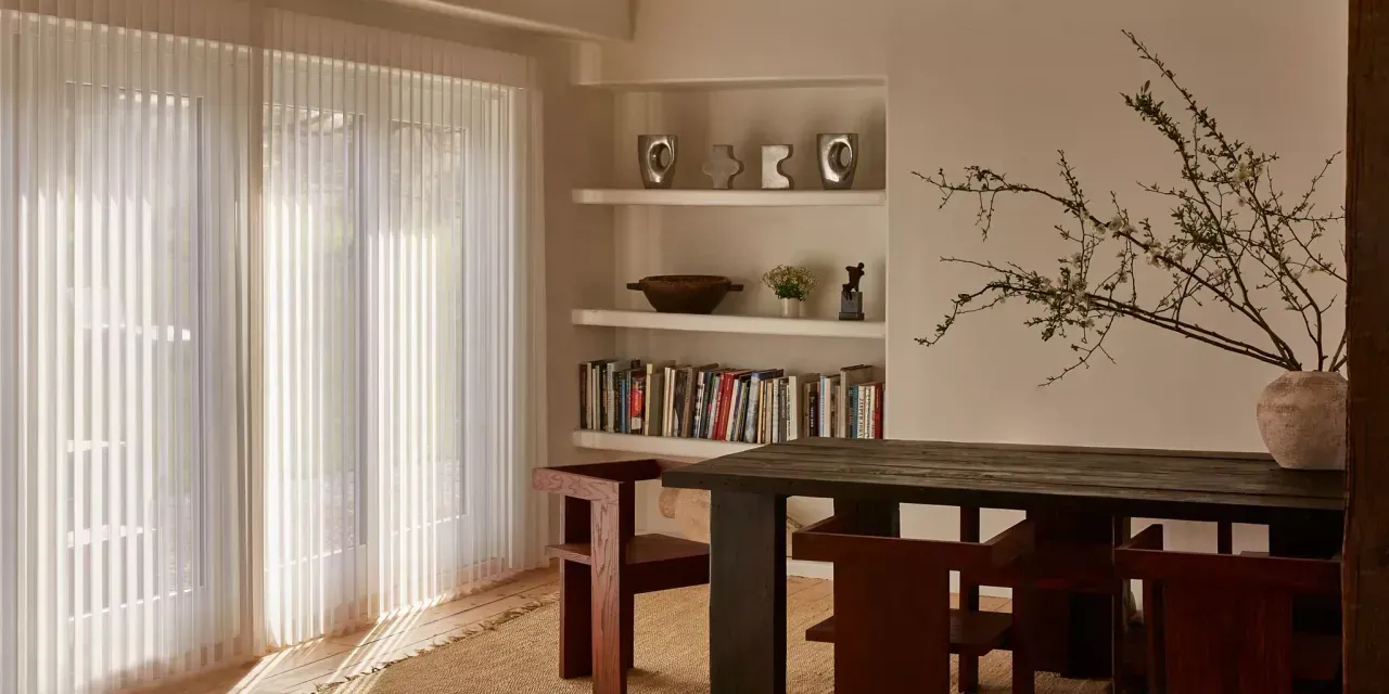 A dining room with a table set with chairs, a bookshelf, and sheer curtains. A vase with branches is on the table.