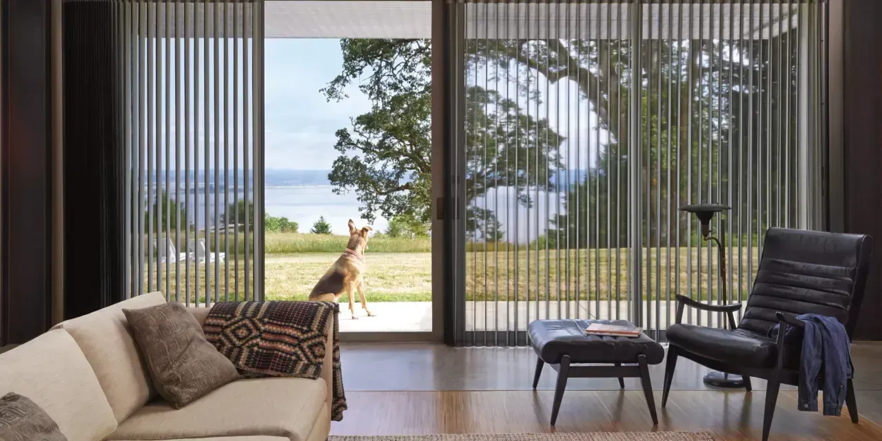 A living room with a large window that looks out to a deer in a field. Beige sofa, black chair, and ottoman.