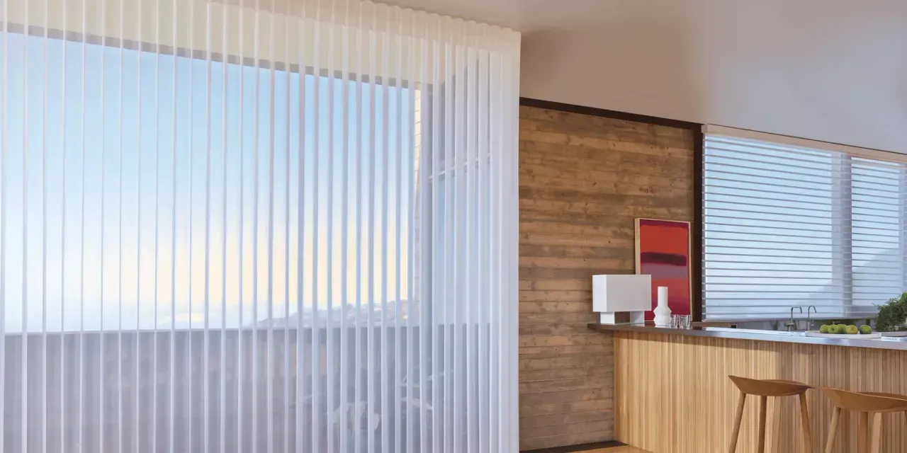 A bright, modern kitchen with white vertical blinds covering a large window.  A wooden accent wall and counter are visible, with bar stools.