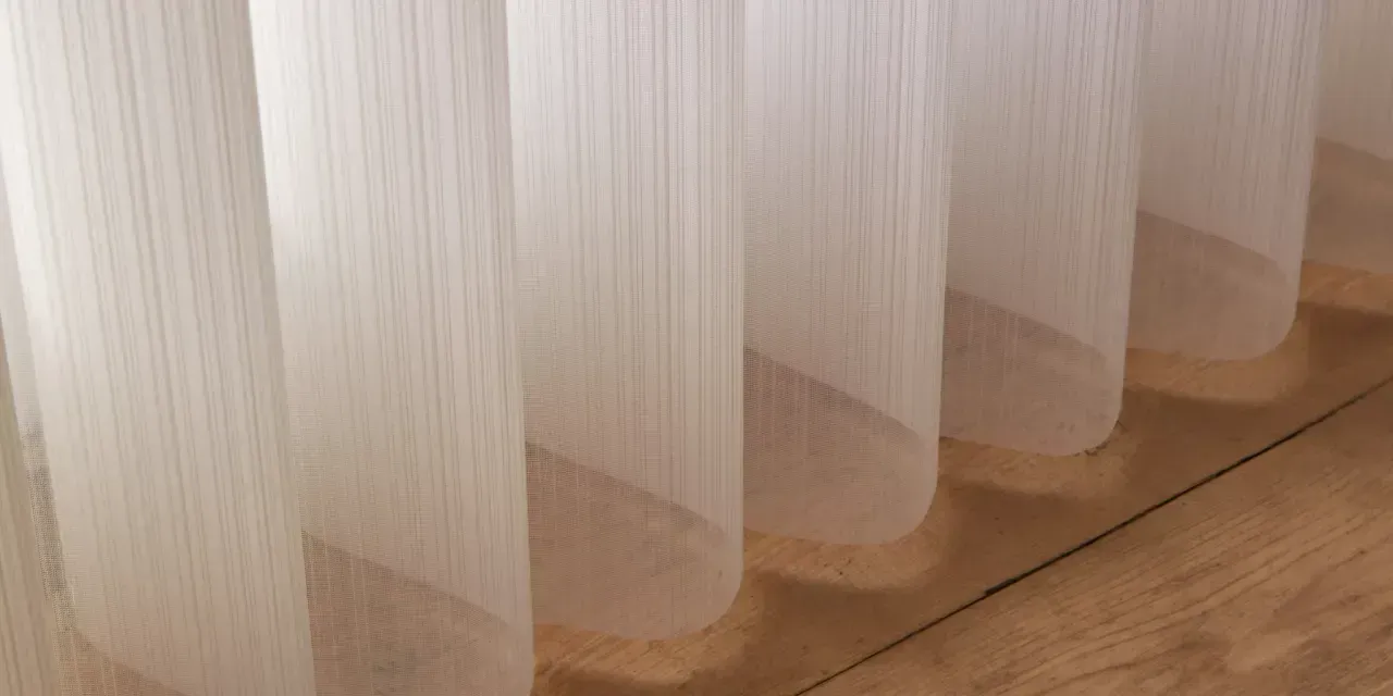 White, sheer curtains hang in vertical folds against a brown wooden floor, catching the light.