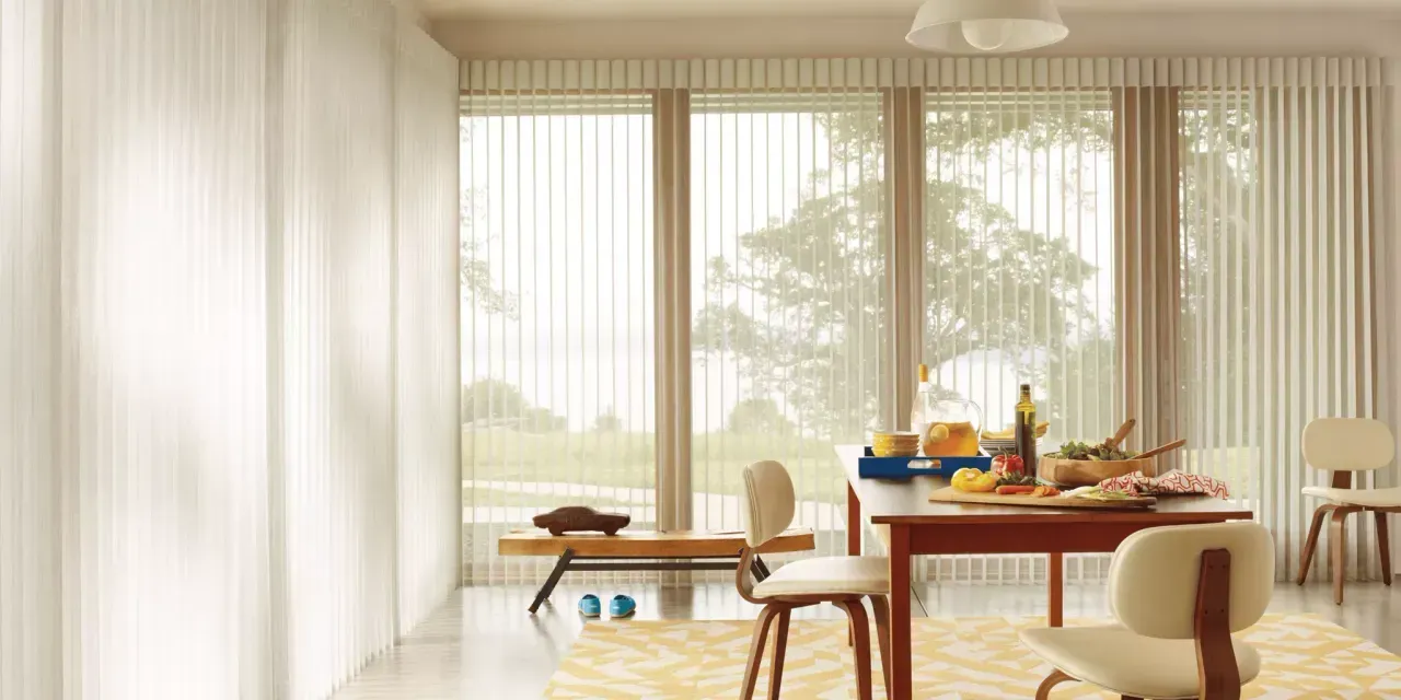 Bright dining room with a table set for a meal, facing large vertical blinds. Beige chairs, rug, and wood accents.