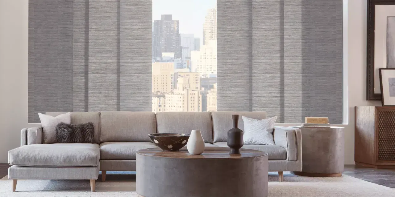 A living room with a grey sectional sofa, round coffee table, and panel shades in front of a window with a city view.