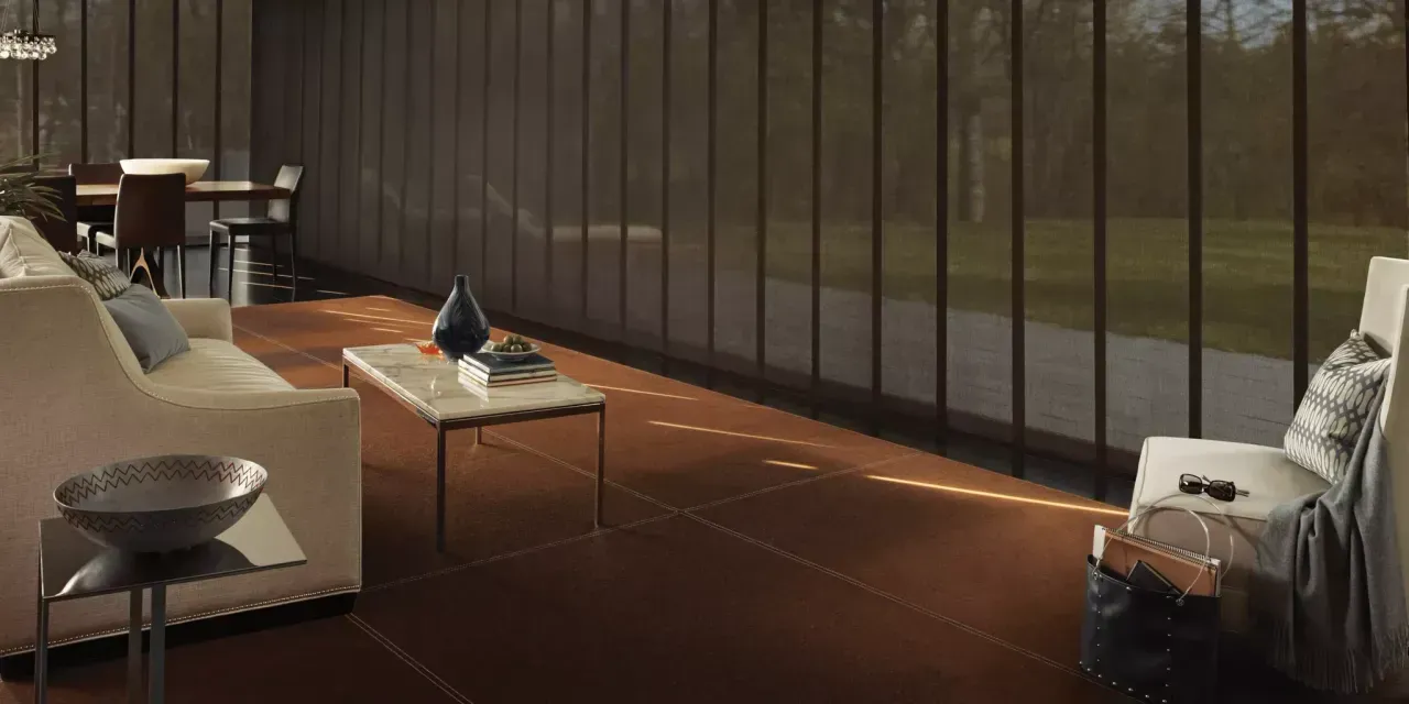 A modern living room with floor-to-ceiling windows and brown panel blinds. The room has a tan sofa, coffee table, and a white armchair.