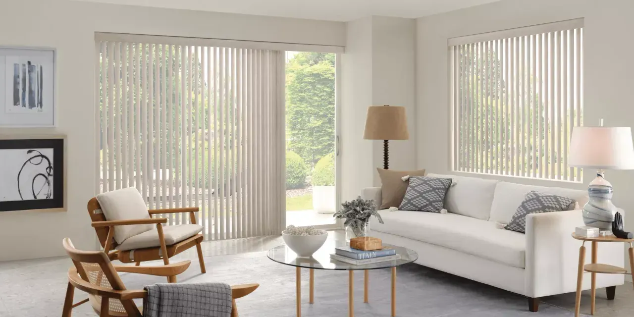 A bright living room with cream vertical blinds, white couch, and wooden armchairs. A large window overlooks a green outdoor space.