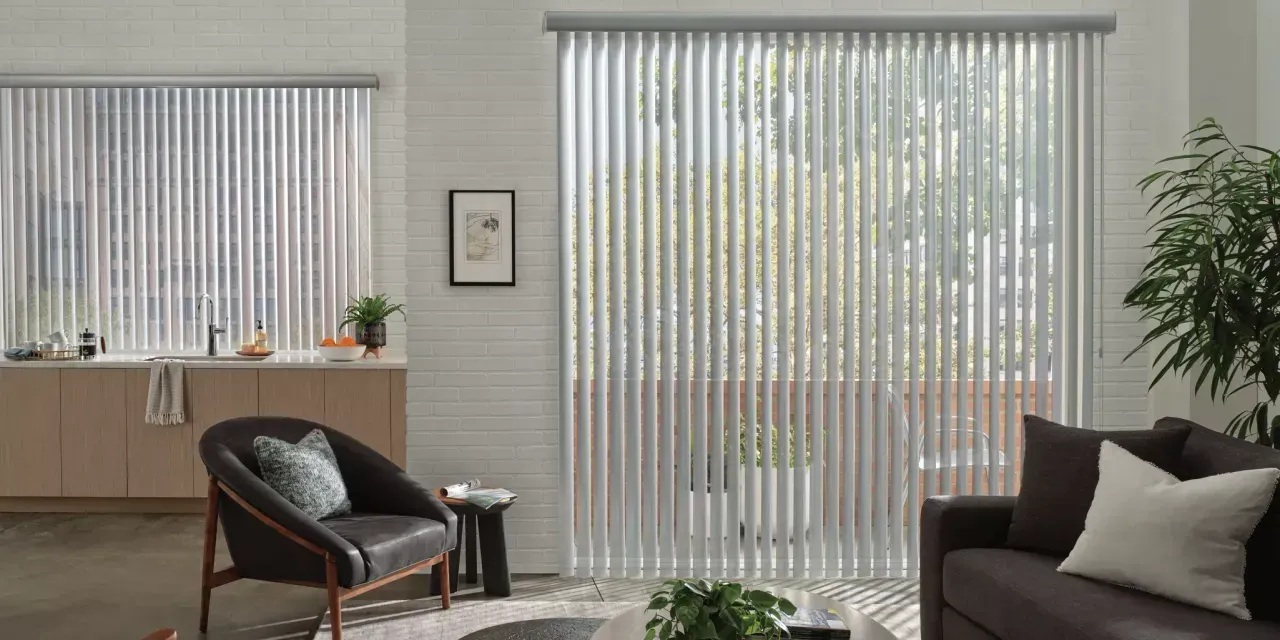 Living room with large vertical blinds, leather chair, and a sofa. Sunlight streams through the window, illuminating the interior.