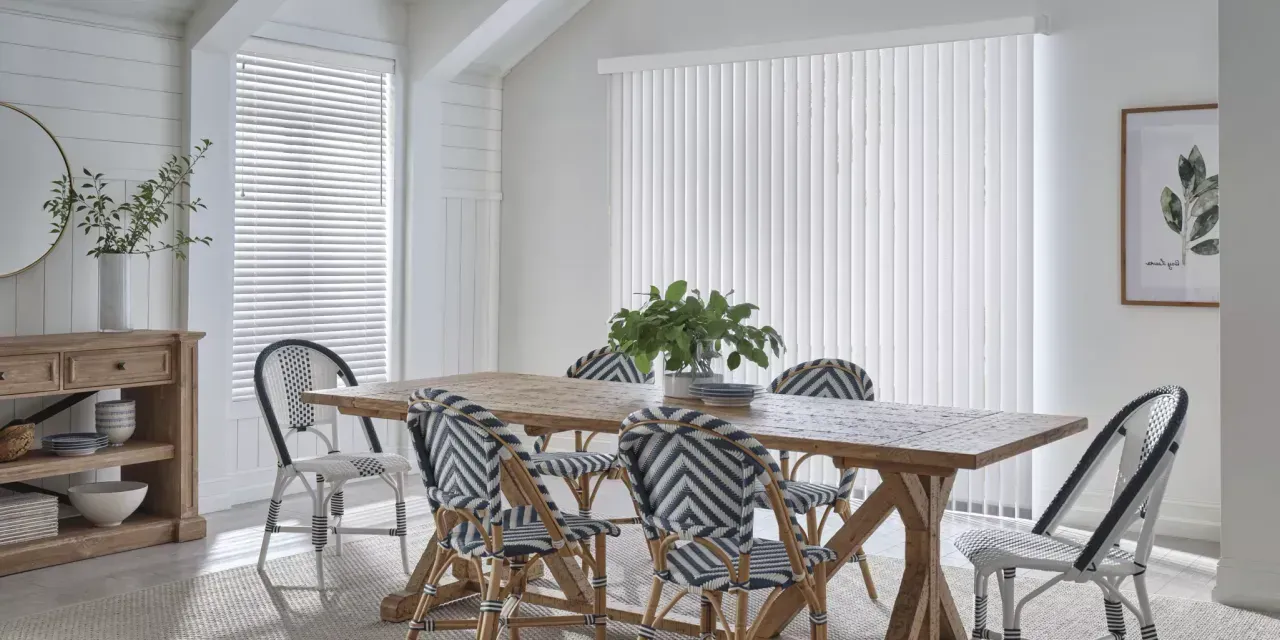 A bright dining room features a wooden table, chairs with black and white chevron-patterned backs, and white blinds, all against white walls.