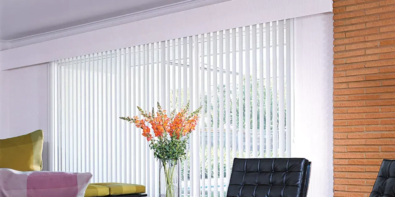 A light-filled interior with white vertical blinds, a vase of flowers, and modern black chairs.  An exposed brick wall is on the right.