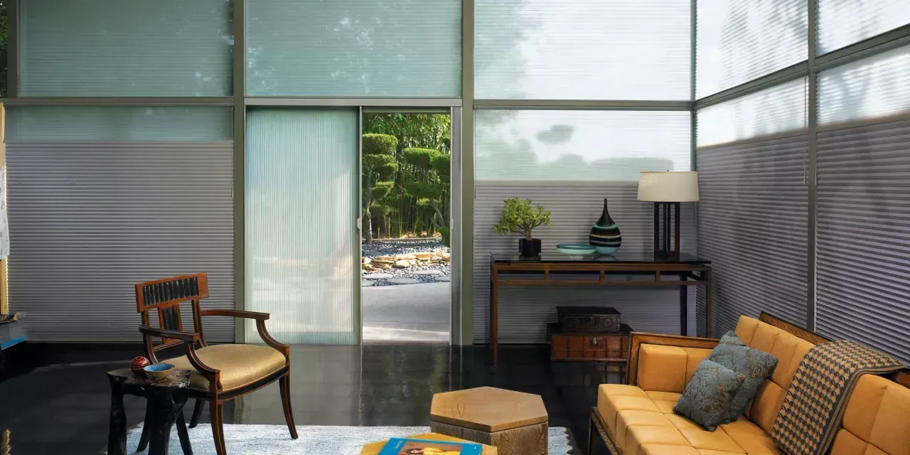 A modern living room with large windows and a view of nature. A wooden sofa, chair, and table are present, along with blinds.