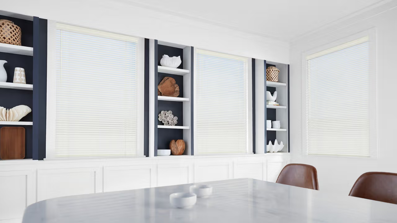 White room with closed blinds and built-in shelves holding decorative items. Brown chairs face a marble table.