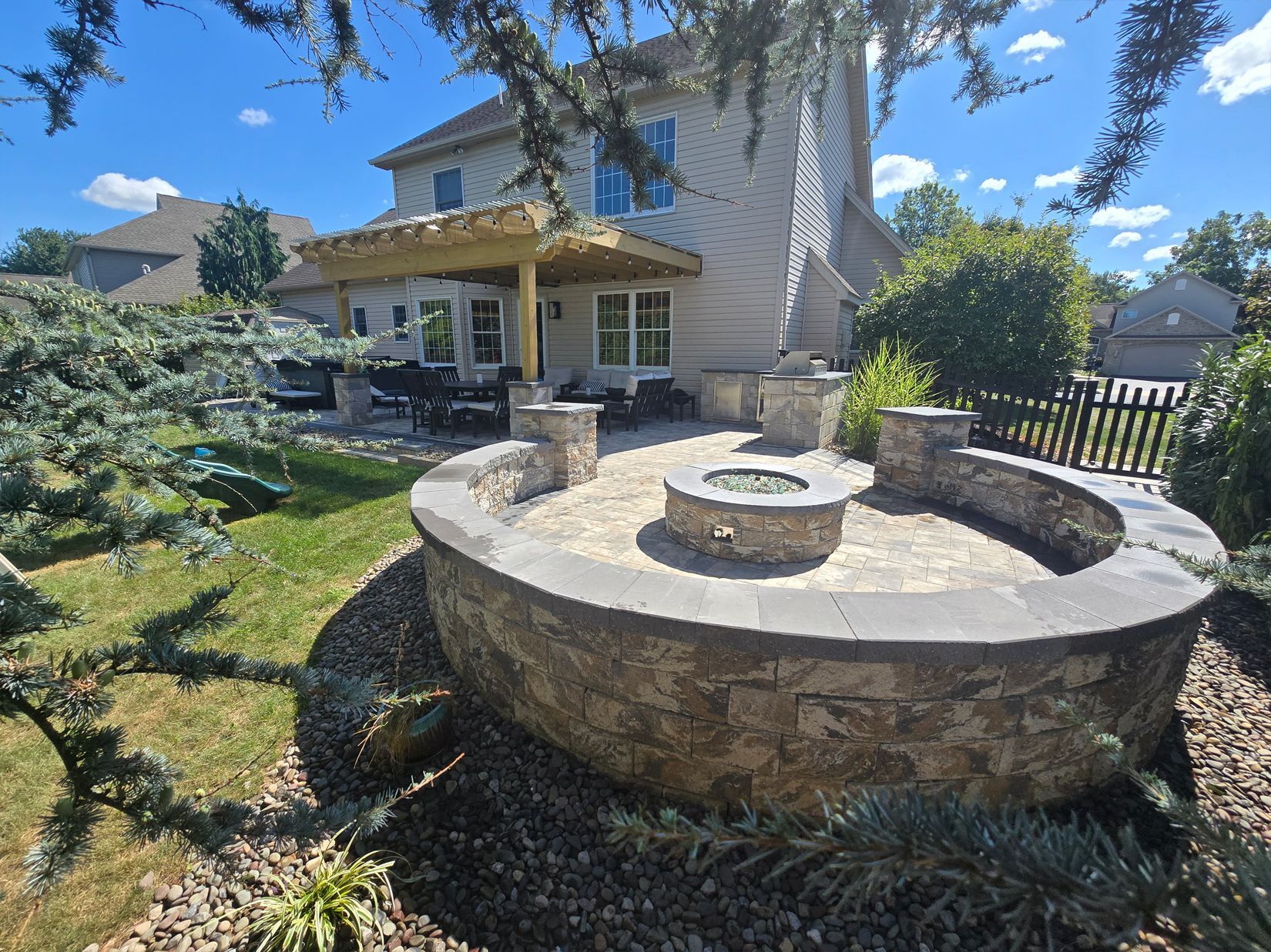 Backyard with a stone fire pit, seating, and covered patio, near a two-story house with blue sky.