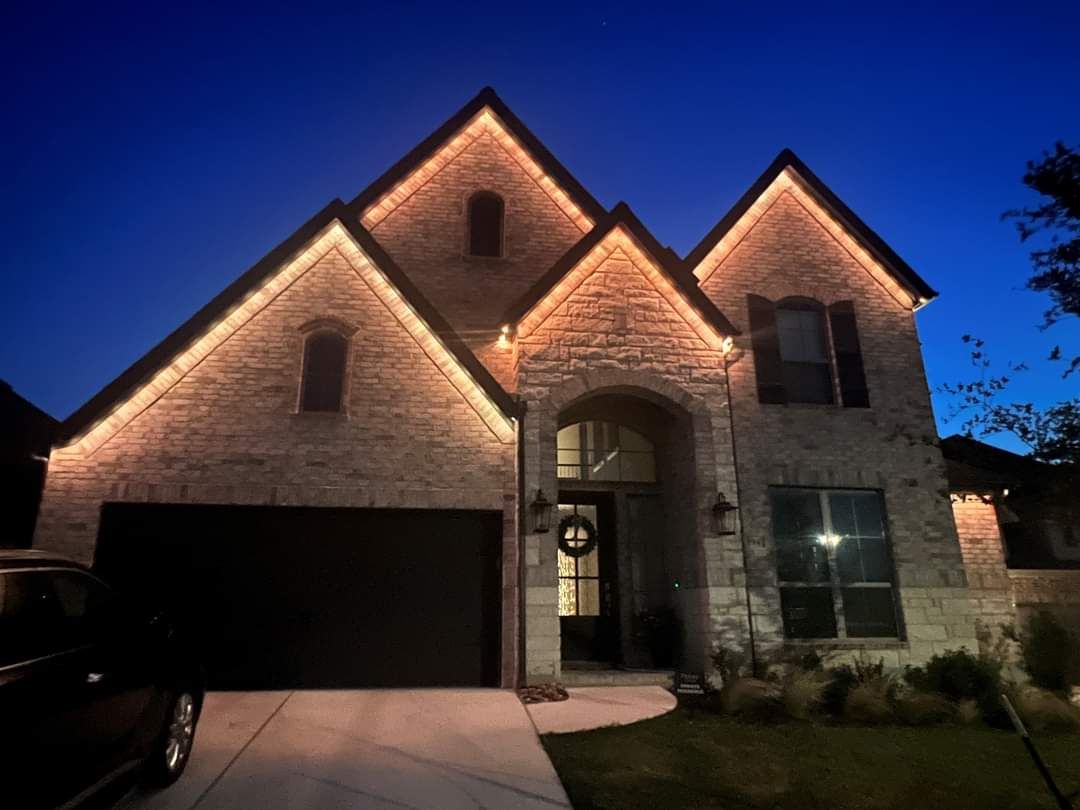 Two-story brick home at dusk, illuminated with warm white lights outlining the roof, a wreath on the front door, and a car in the driveway.