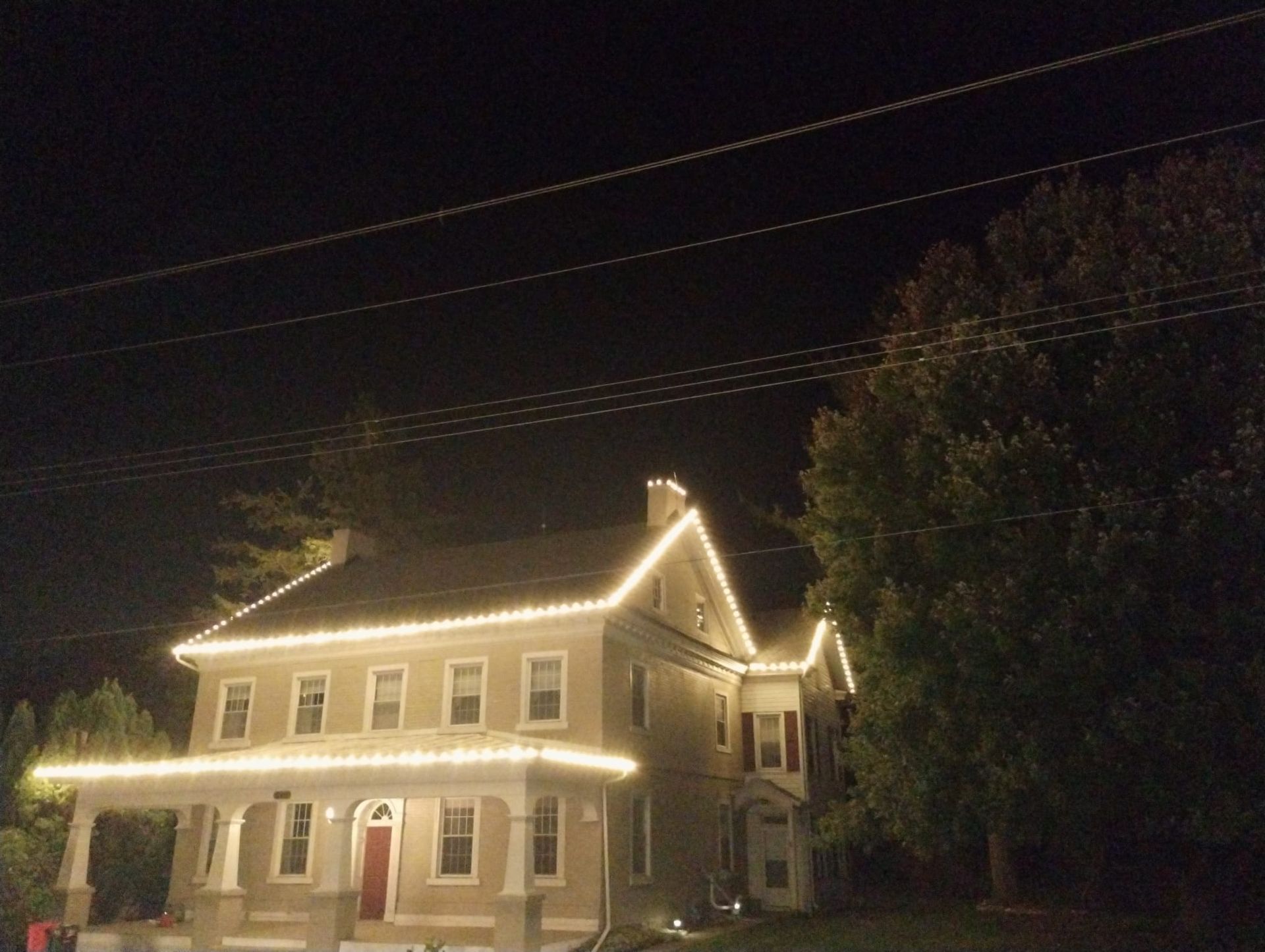 A two-story house at night, lit with white lights along the roofline and porch; wires above.