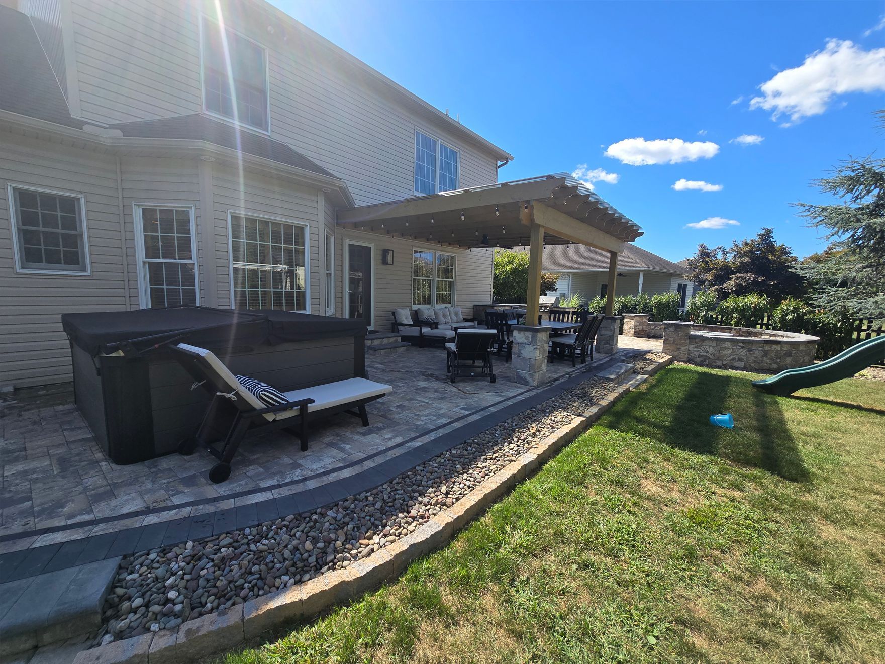Backyard patio with hot tub, pergola, and seating under a sunny, blue sky.
