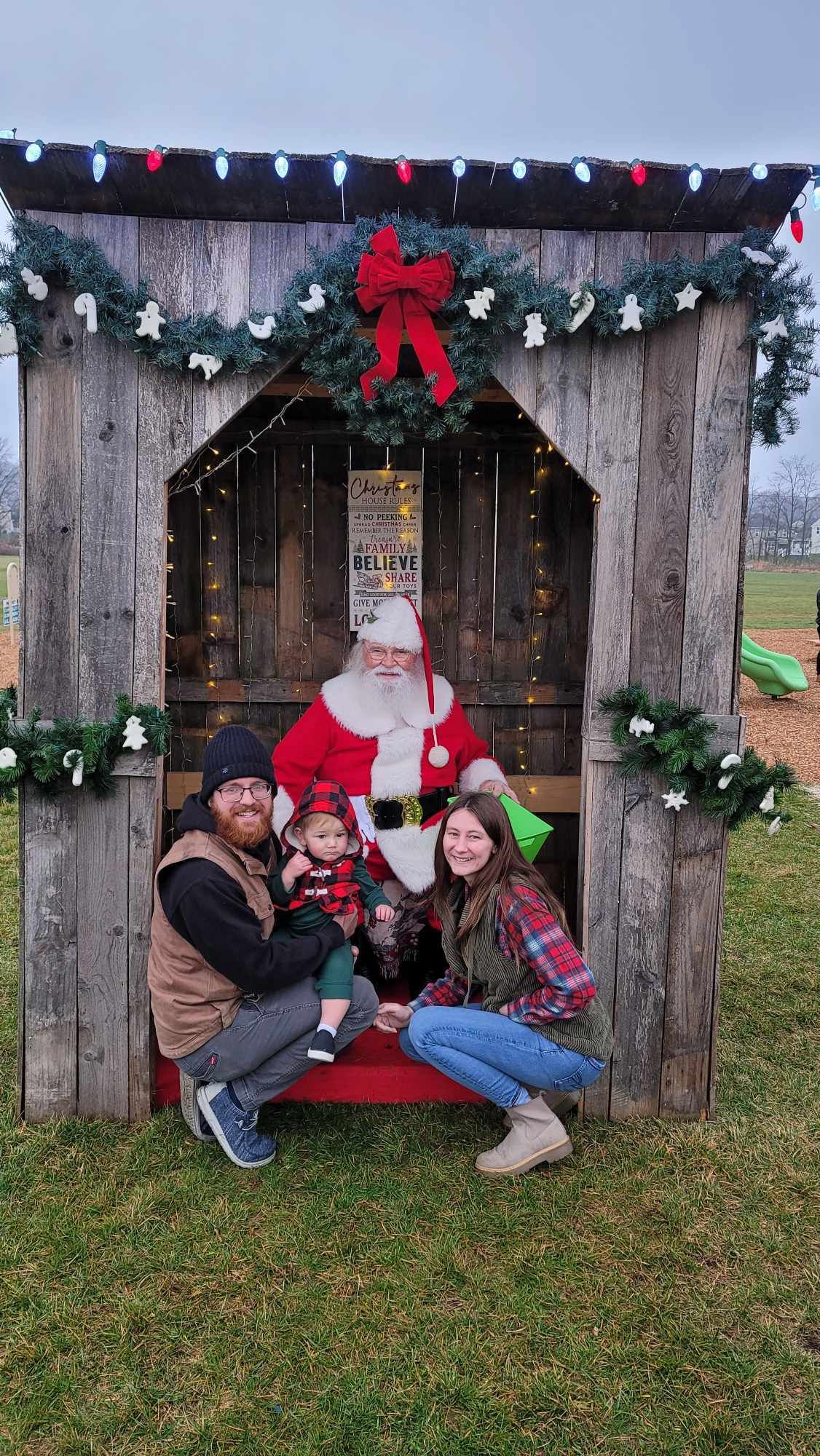 Family with Santa in a wooden booth. Parents flank Santa with toddler. Holiday decorations.