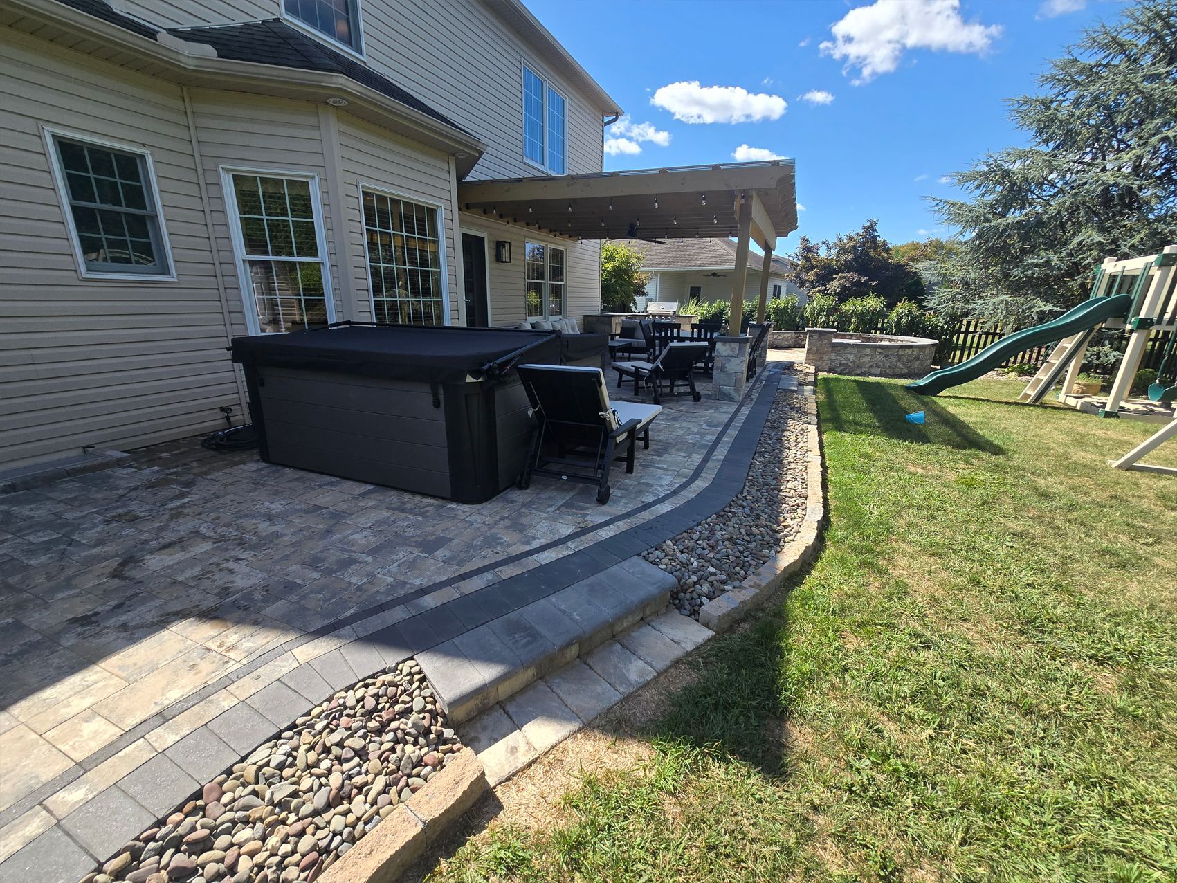 Backyard patio with hot tub, seating, and covered area. Green grass, blue sky, and playground visible.