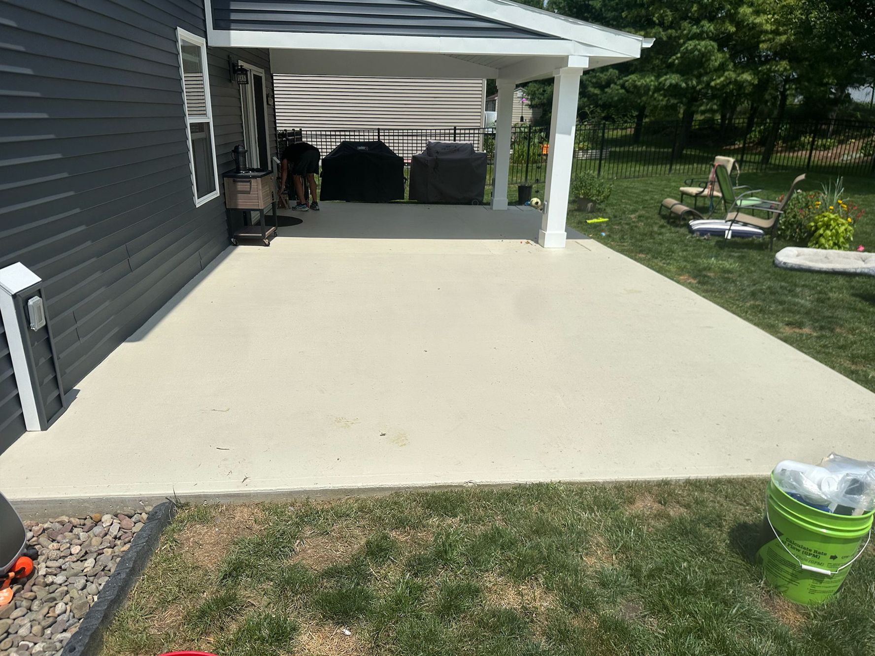 A concrete patio extends from a dark gray house, covered porch has white supports. Green grass surrounds it.