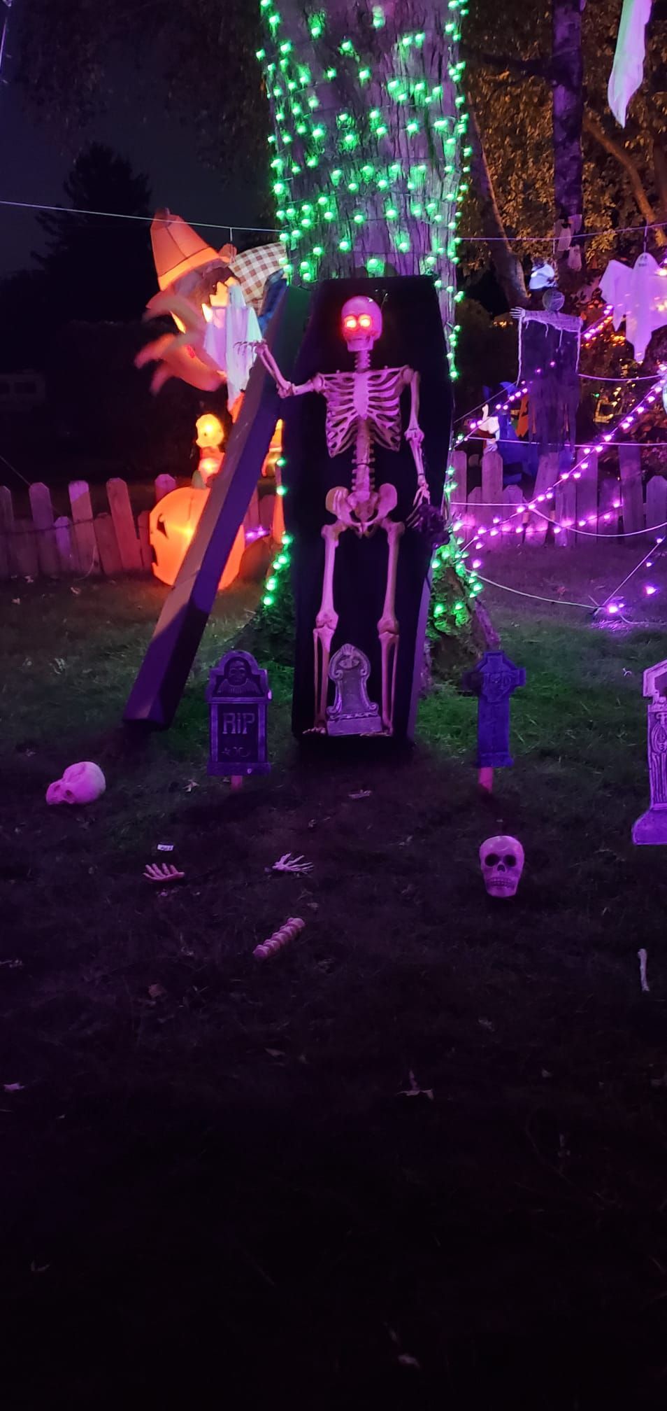A Halloween display featuring a skeleton, tombstones, and a witch, illuminated with green and purple lights.