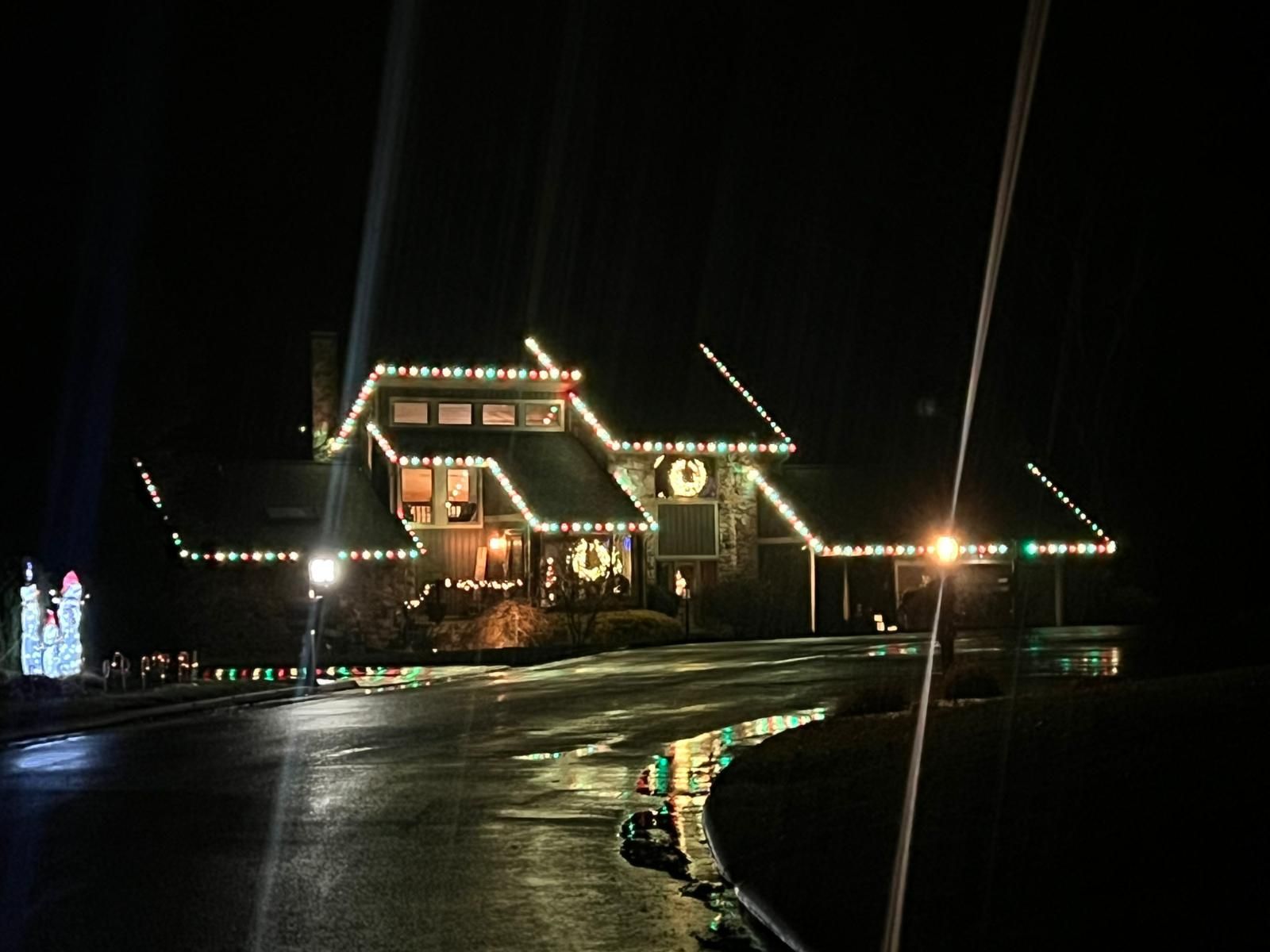House at night, decorated with Christmas lights and wreaths; wet driveway reflects lights.