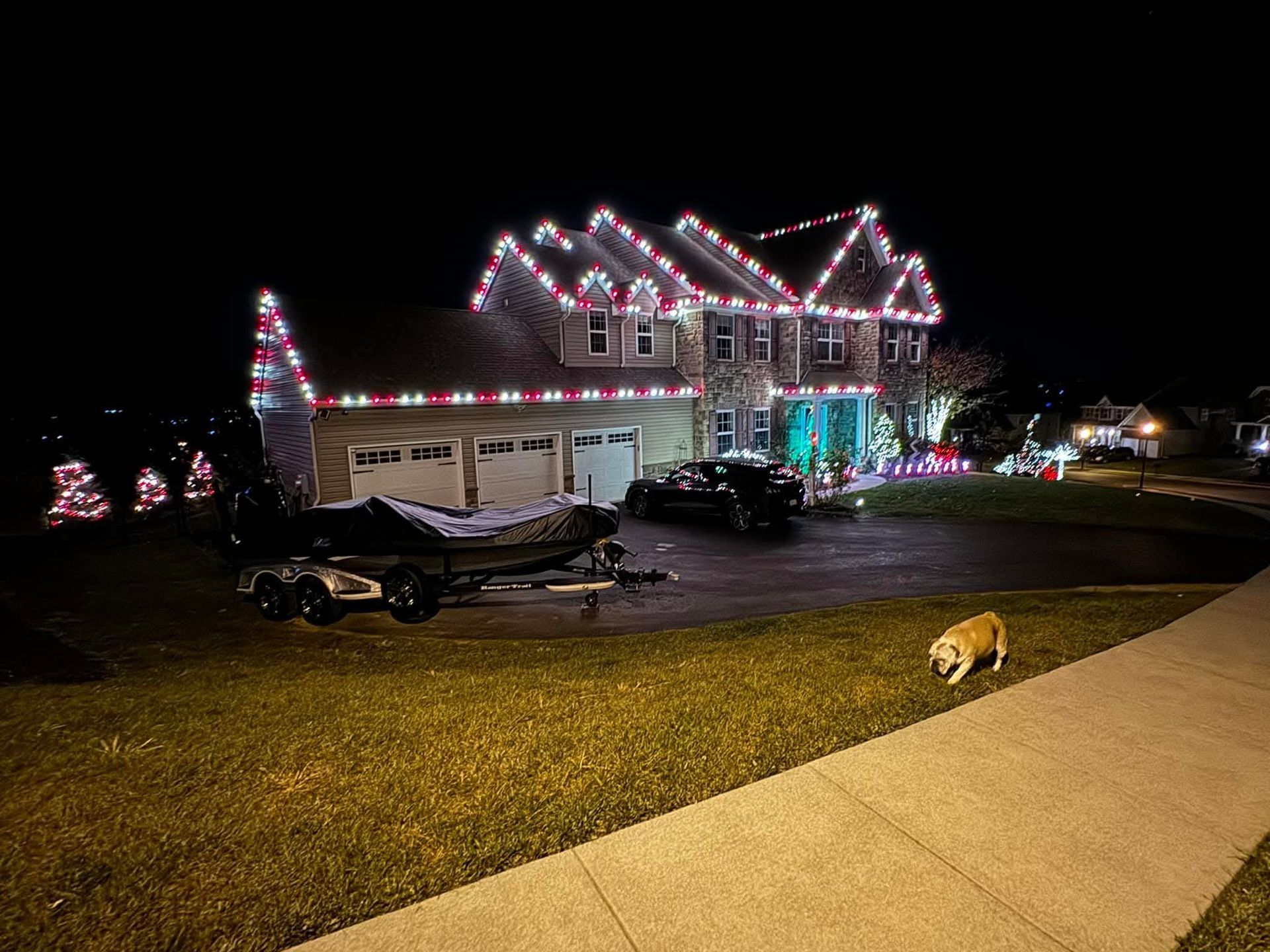 House decorated with red and green Christmas lights, boat in driveway, dog on lawn.