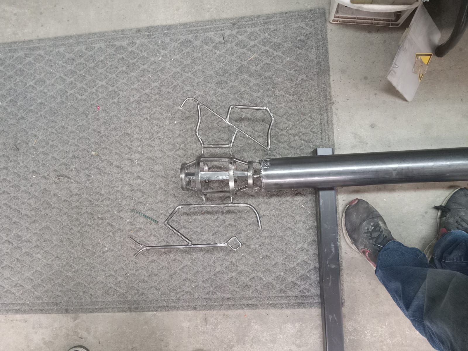 Metal parts and tools on a gray mat in a workshop. A long, dark pipe is in the frame.