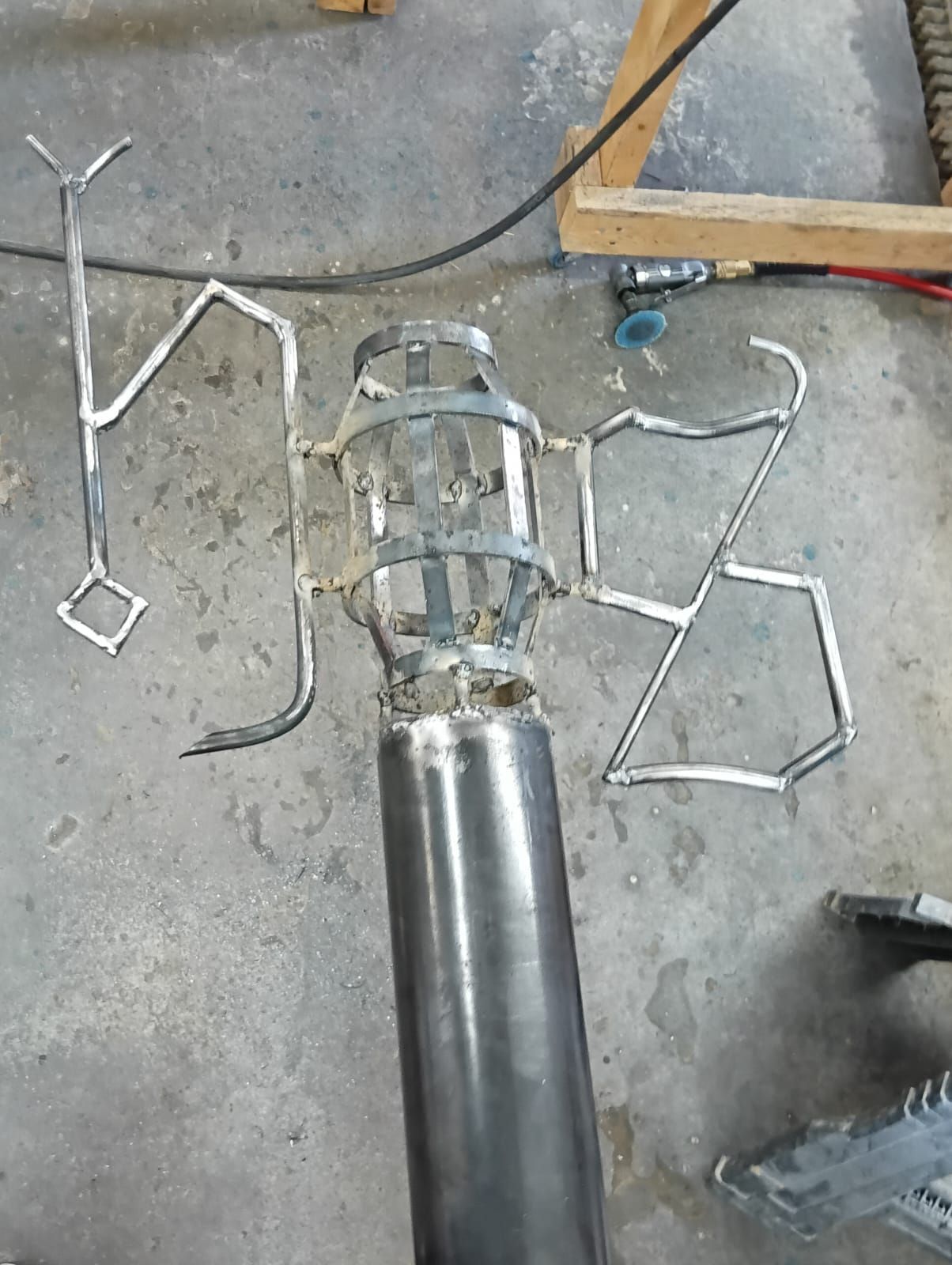 Welded metal sculpture in progress on a concrete floor. A cylindrical form is topped with abstract arms and shapes.