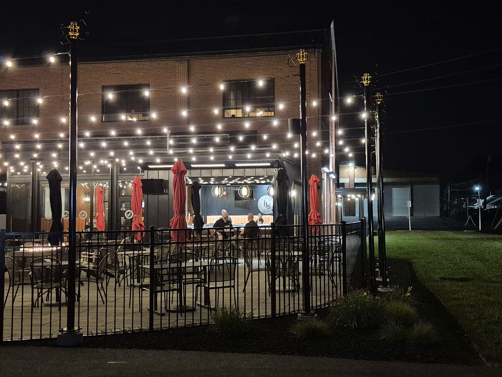 Night view of a restaurant patio with string lights, brick building, red umbrellas, and black metal fence.