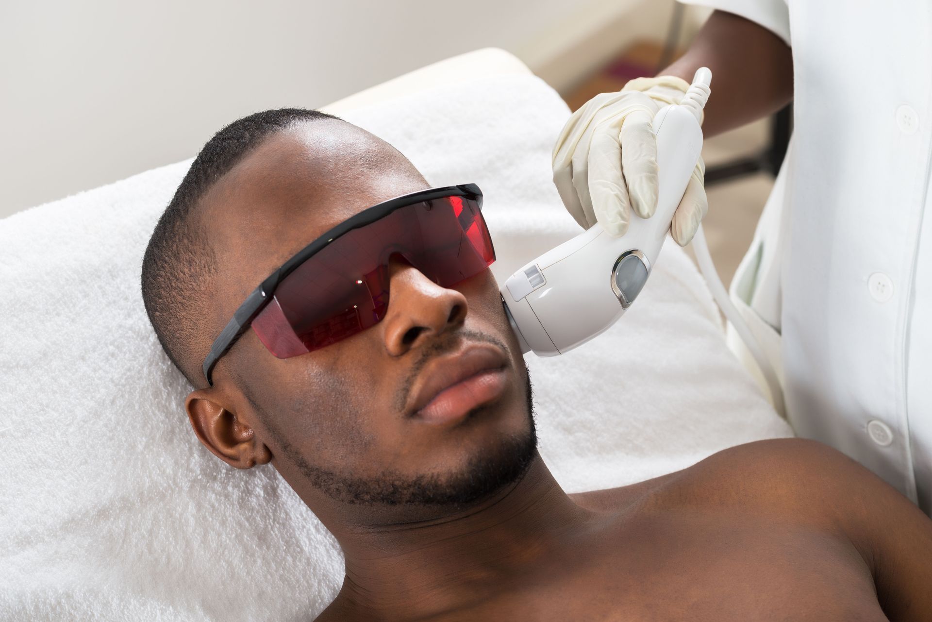 Man receiving laser hair removal treatment on his face, wearing safety glasses.