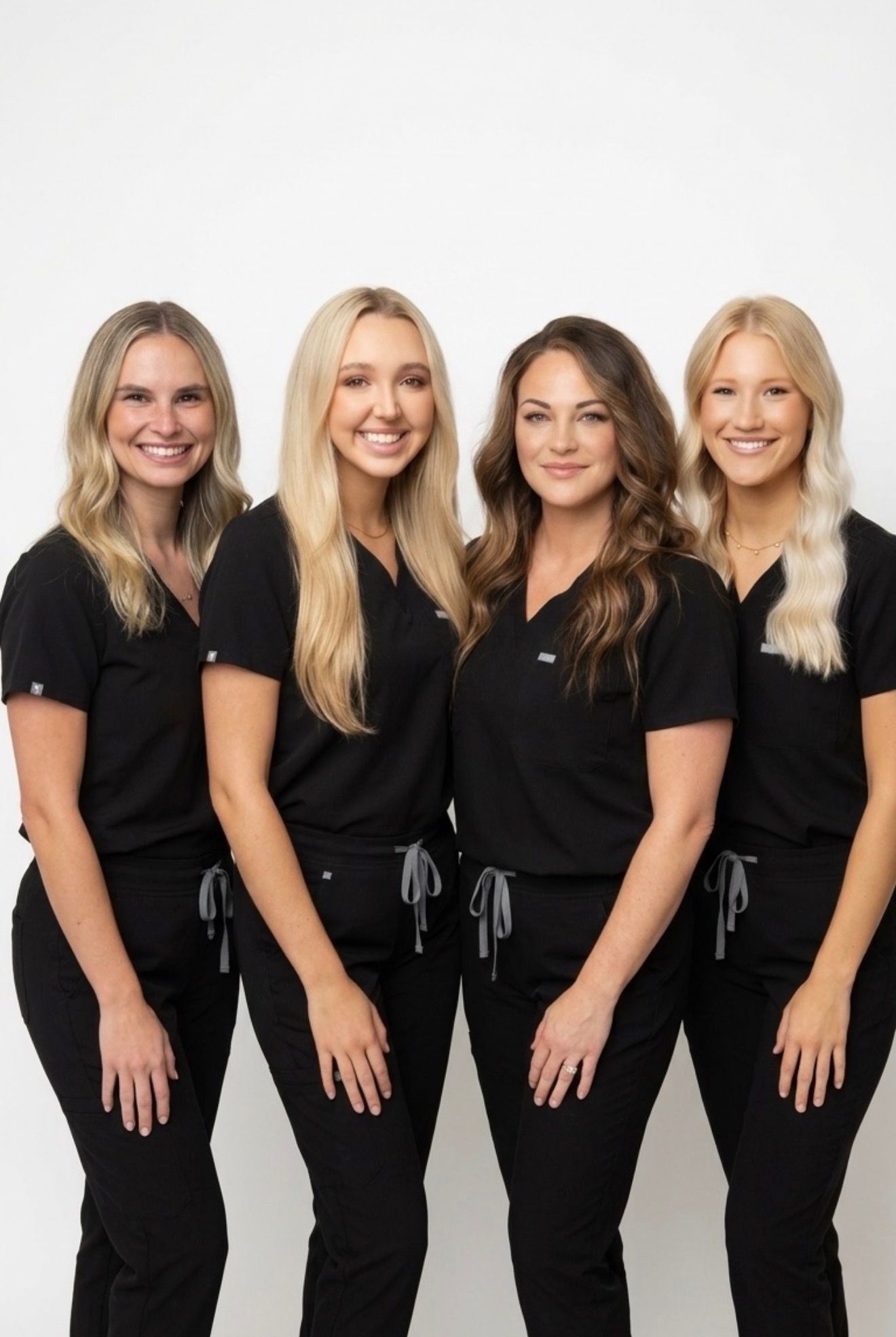 Four women in black outfits standing together against a white background, smiling at the camera