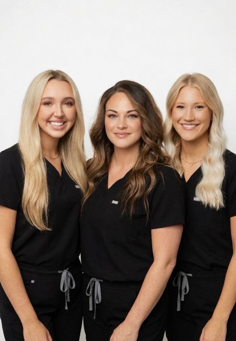 Three women in black outfits standing side by side against a white background, smiling.