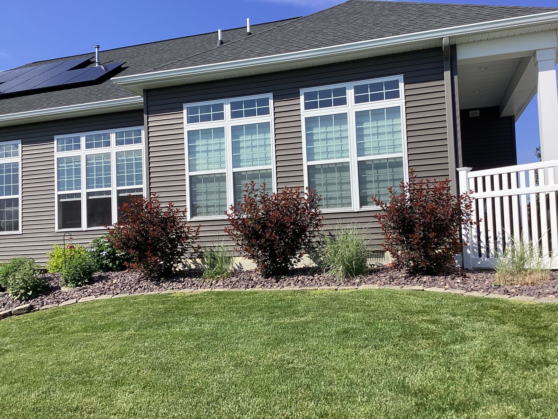 House exterior with gray siding, white-framed windows, and landscaping featuring red bushes and green lawn.