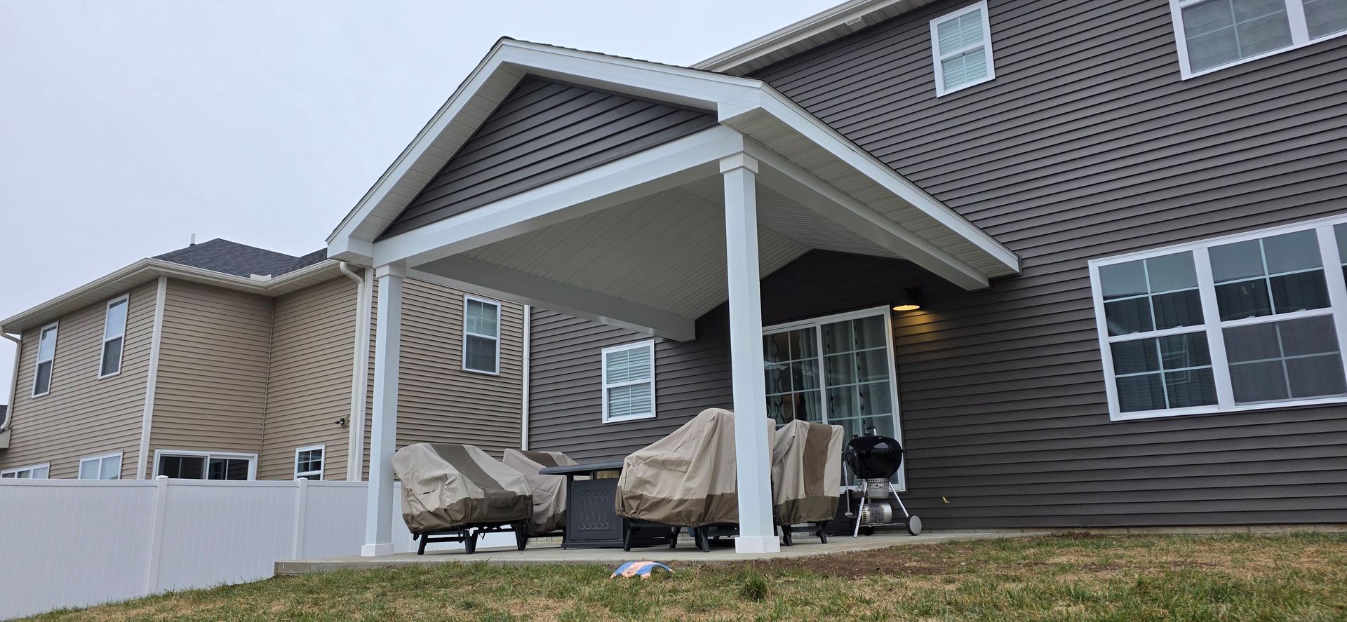 Two covered patio chairs under a white awning beside a gray house with large windows