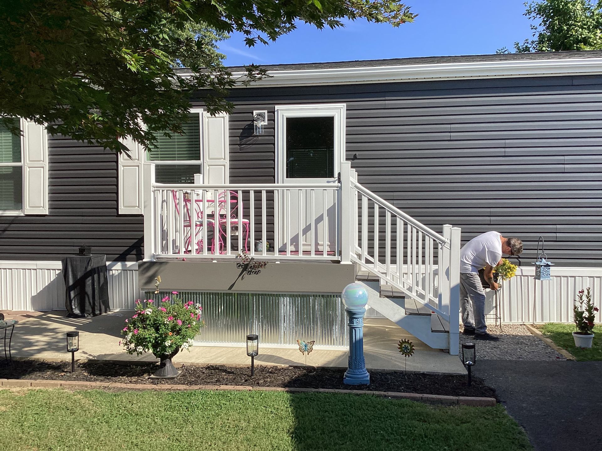 Exterior of a dark gray mobile home with a white porch and steps; a person is near the steps, tending to flowers.