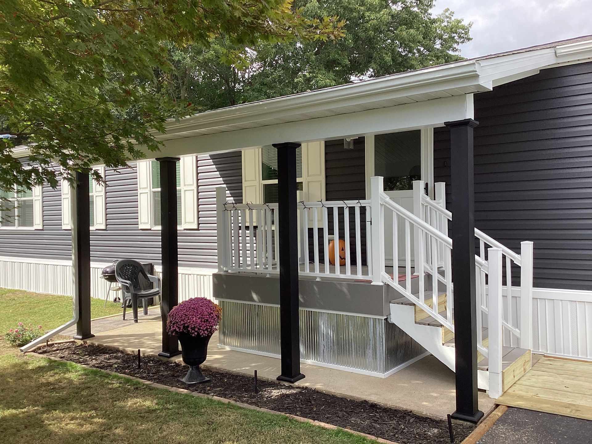 Porch of a gray house with a white railing and black columns. Pink flowers in a black pot.