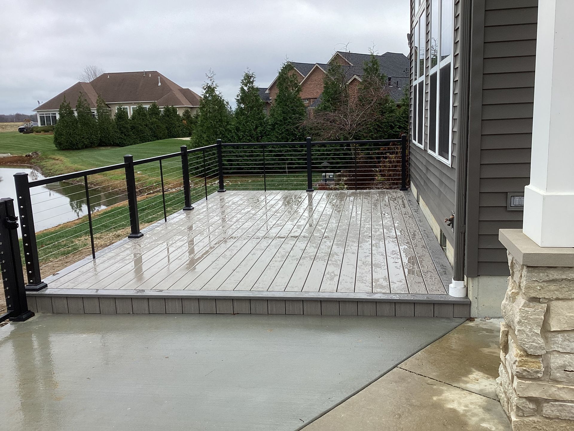 Wet, gray composite deck with black railing next to a pond. House in the background. Overcast day.