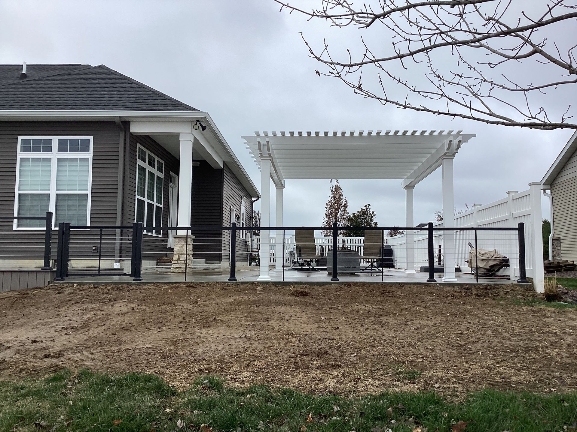 Outdoor deck with white pergola, black metal railing, adjacent to a house with dark siding. Cloudy sky.