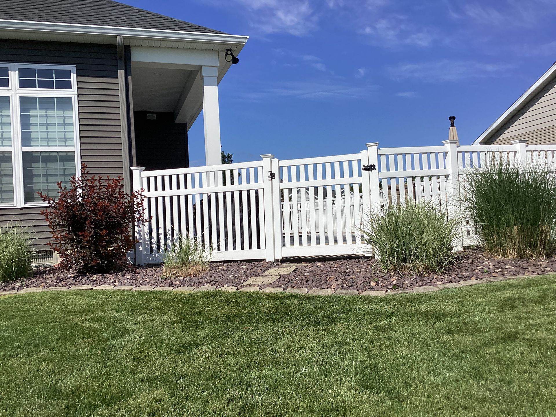 White picket fence with gate, bordering a house with porch, shrubs, and grass lawn under blue sky.