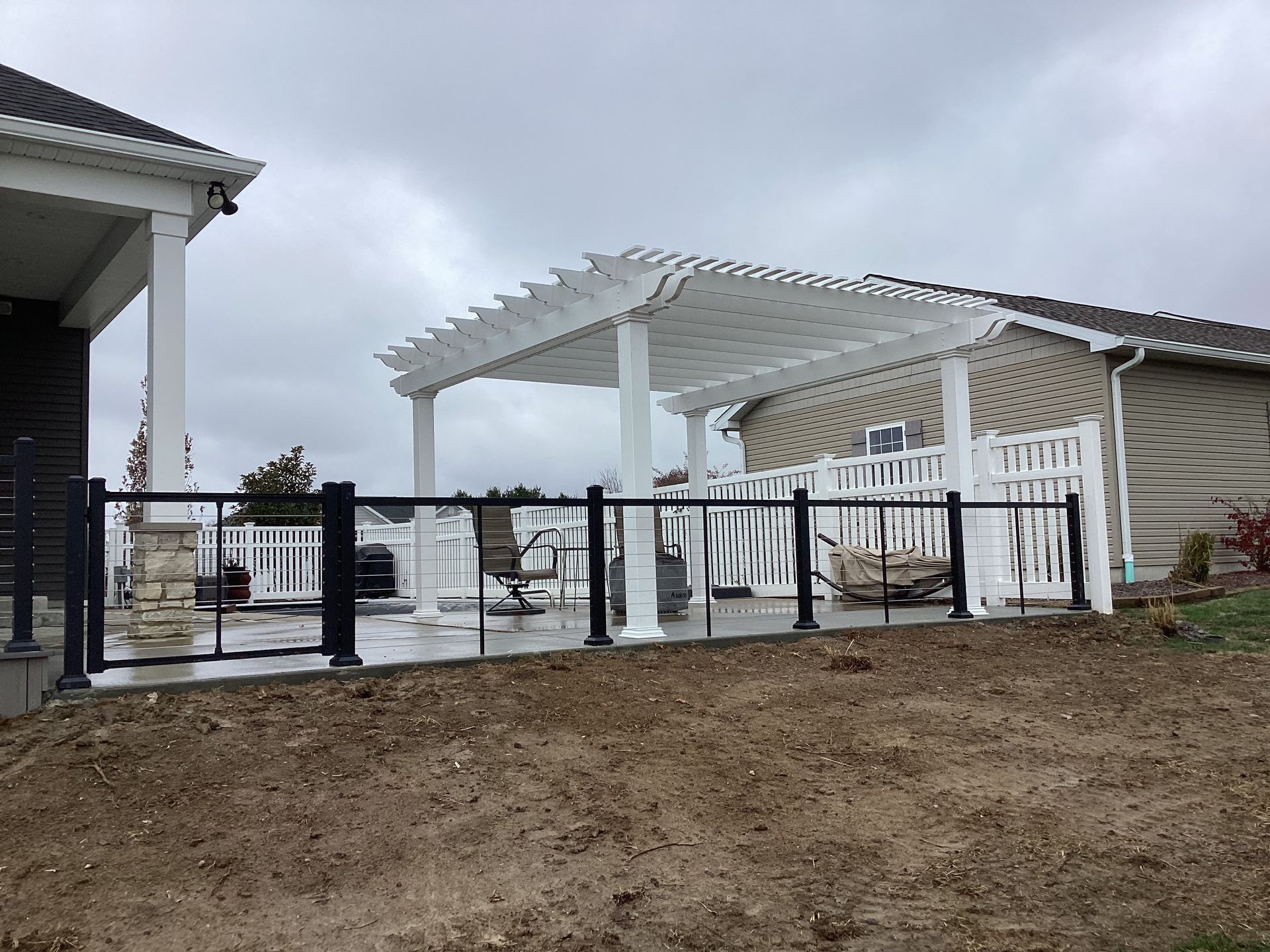 White pergola patio with black fence and railings beside a tan house on a cloudy day
