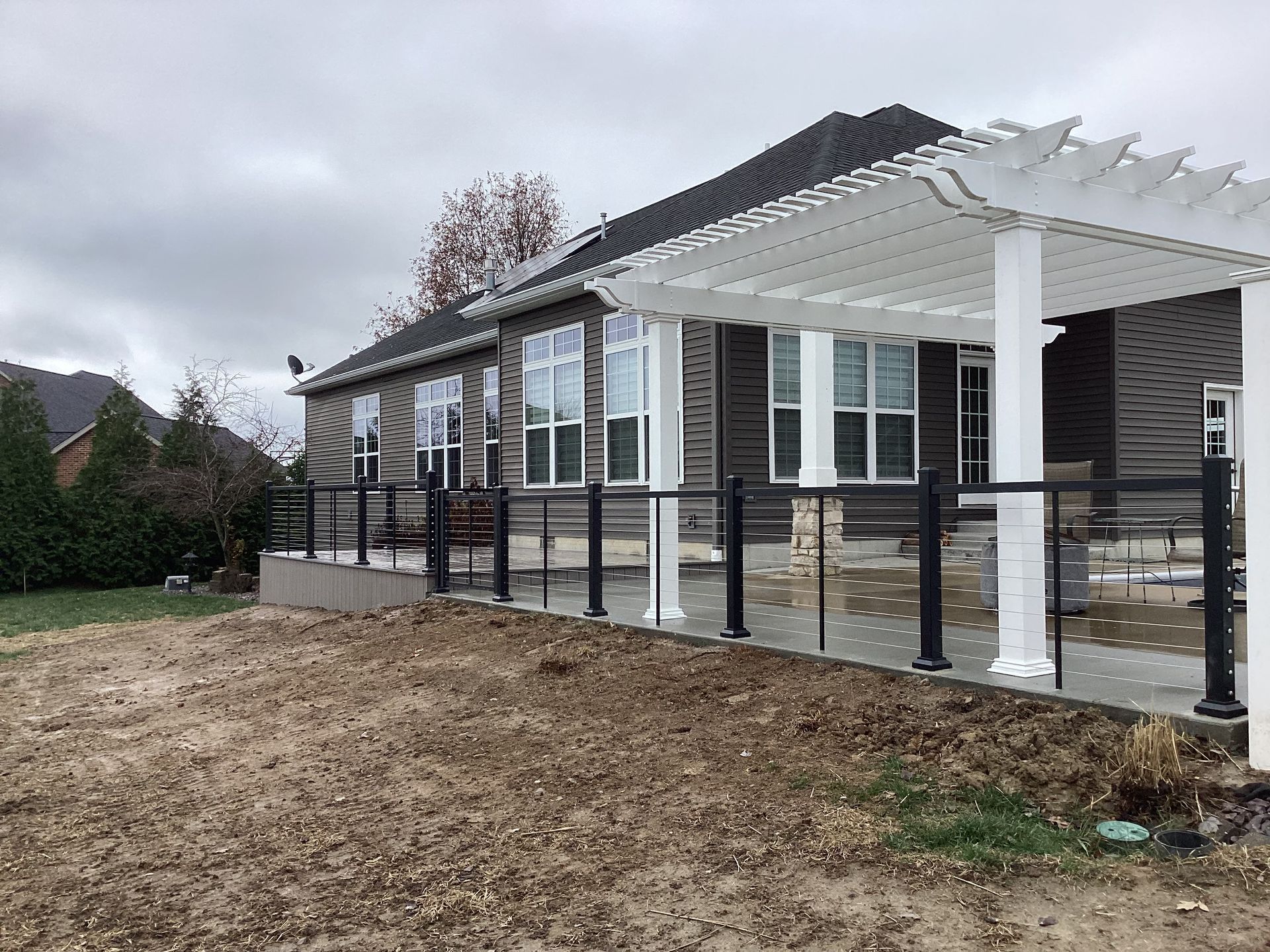 House with a white pergola and black railing on a bare yard under cloudy skies