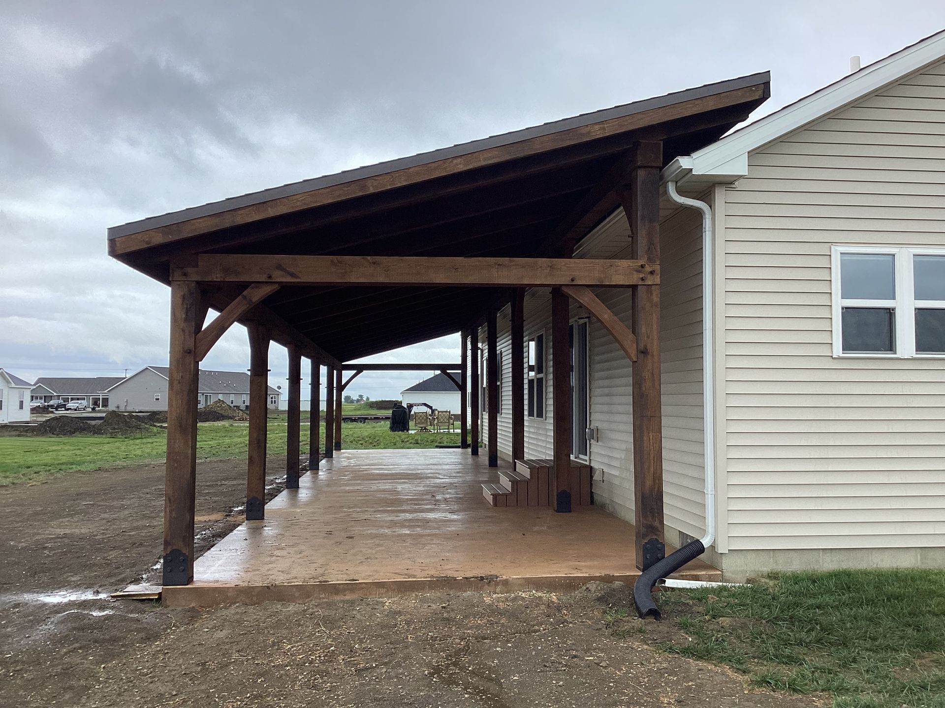 Covered wooden walkway attached to a beige house, with wet concrete and open yard beyond.