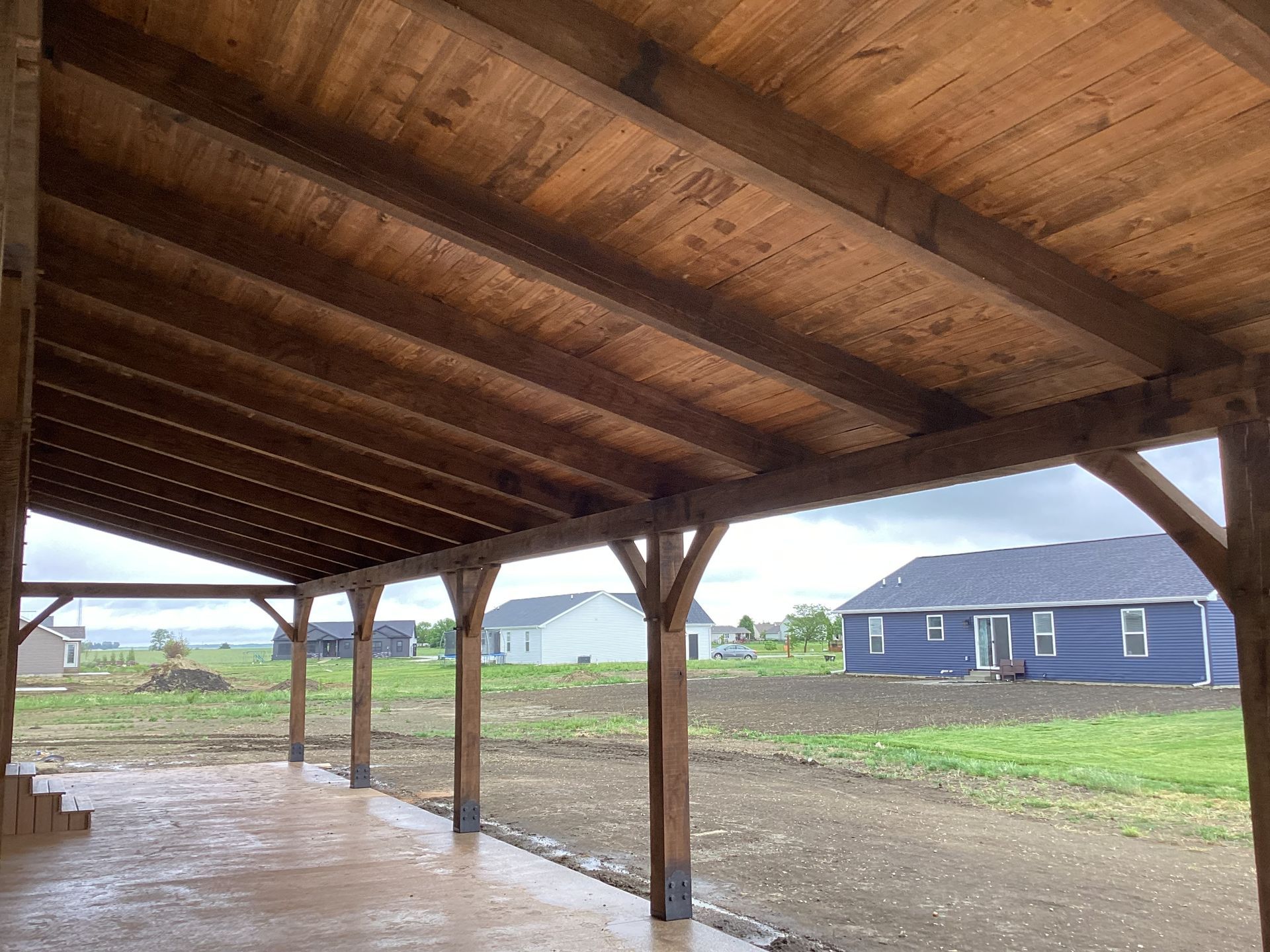 Wooden carport with open sides on a wet concrete slab, overlooking a grassy rural lot and blue house.