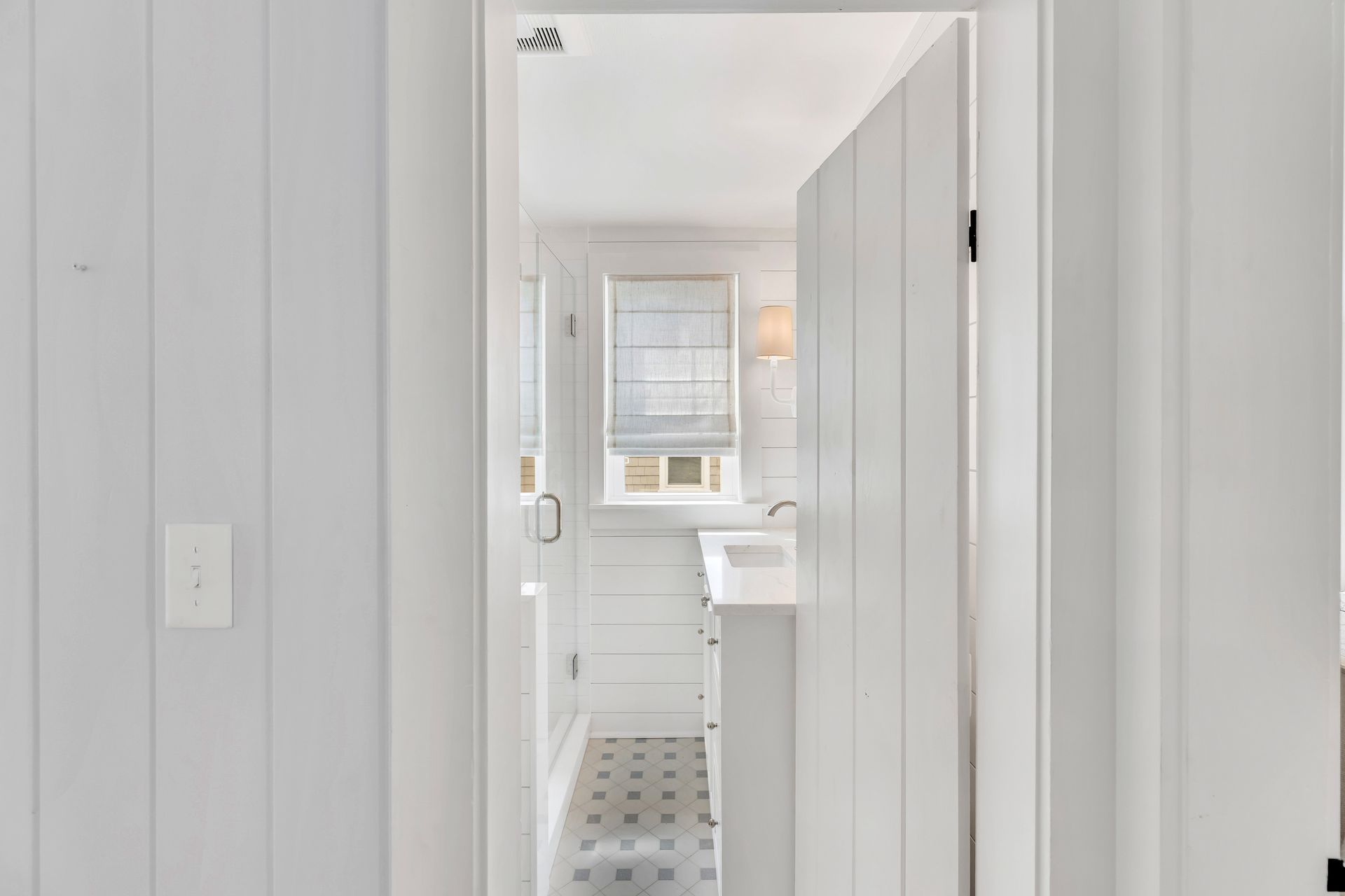 View through a white doorway into a bright bathroom with a sink, window, and patterned tile floor